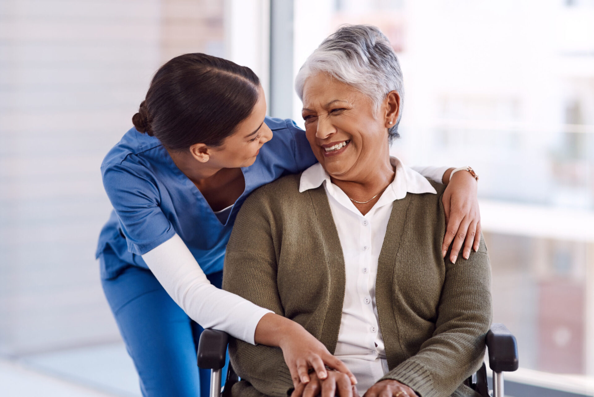 Healthcare, disability and a nurse hugging an old woman in a wheelchair during a nursing home visit. Medical, hug and funny with a laughing female medicine professional talking to a senior resident